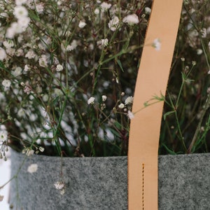 May include: A gray felt planter with a light brown leather handle. The planter is filled with white baby's breath flowers.
