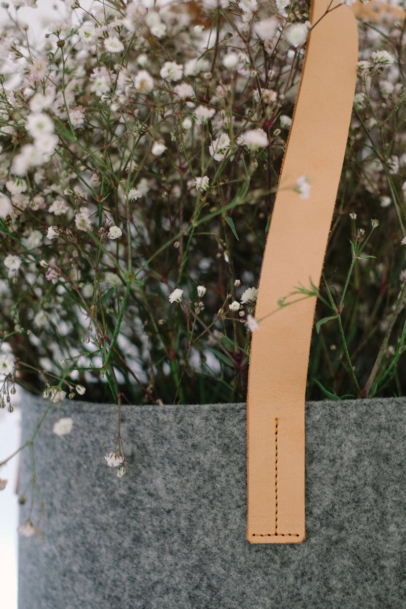 May include: A gray felt planter with a light brown leather handle. The planter is filled with white baby's breath flowers.