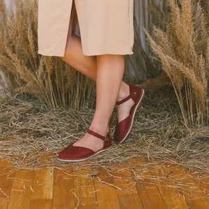 Pu&ograve; includere: Un paio di sandali in pelle color borgogna con cinturini alla caviglia. Le sandali sono indossate da una persona in piedi su un pavimento di legno con erba secca sullo sfondo.