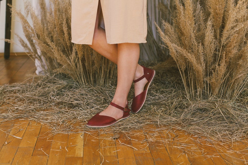 Pu&ograve; includere: Un paio di sandali in pelle color borgogna con cinturini alla caviglia. Le sandali sono indossate da una persona in piedi su un pavimento di legno con erba secca sullo sfondo.