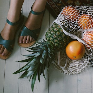 May include: A white mesh bag filled with oranges and a pineapple. The bag is on a wooden floor with a pair of teal blue sandals next to it.