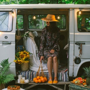 May include: A woman wearing a straw hat sits in a vintage van with string lights. She is holding a net bag filled with oranges. The van is parked in a wooded area with a picnic blanket and a wooden crate.