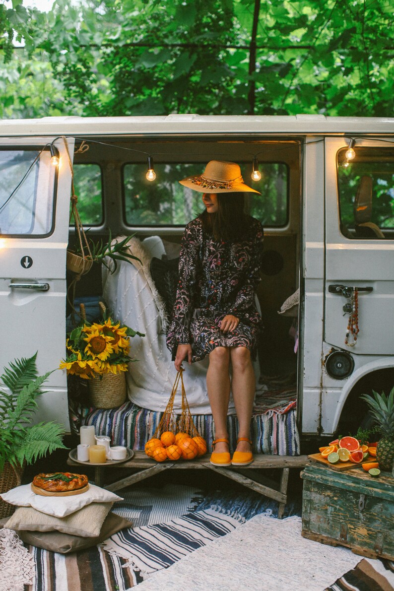 May include: A woman wearing a straw hat sits in a vintage van with string lights. She is holding a net bag filled with oranges. The van is parked in a wooded area with a picnic blanket and a wooden crate.