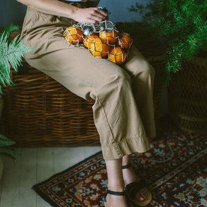 K&ouml;nnte beinhalten: Eine Frau in einem wei&szlig;en Oberteil und braunen Hosen sitzt auf einer Korbbank und h&auml;lt eine Netz-Tasche mit Orangen. Sie tr&auml;gt schwarze Sandalen und sitzt auf einem Teppich mit Blumenmuster.