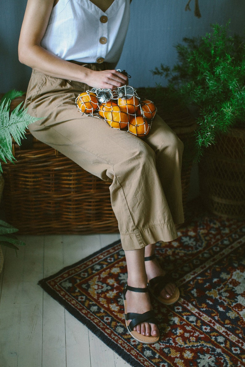 K&ouml;nnte beinhalten: Eine Frau in einem wei&szlig;en Oberteil und braunen Hosen sitzt auf einer Korbbank und h&auml;lt eine Netz-Tasche mit Orangen. Sie tr&auml;gt schwarze Sandalen und sitzt auf einem Teppich mit Blumenmuster.