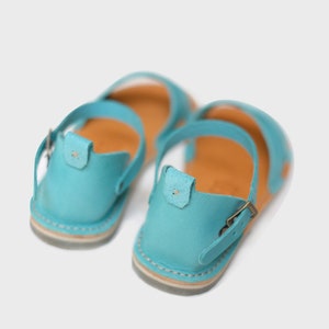 May include: A pair of light blue leather sandals with a buckle closure. The sandals have a brown leather insole and a white sole.