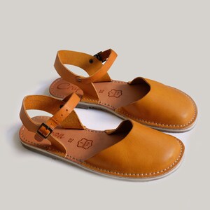 May include: A pair of tan leather sandals with ankle straps and buckles. The sandals have a simple design and are made of high-quality leather.