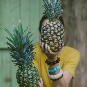 May include: A person holding a green pineapple in front of their face. A second pineapple is sitting on a wooden surface behind them.