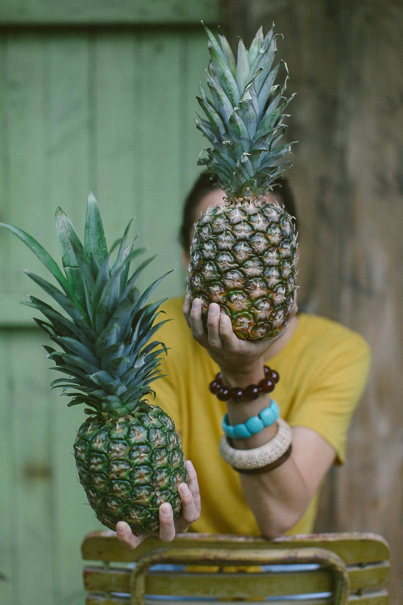 May include: A person holding a green pineapple in front of their face. A second pineapple is sitting on a wooden surface behind them.