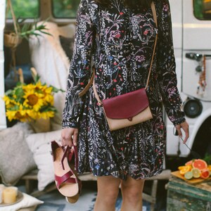 May include: A woman wearing a floral print dress and a burgundy and beige crossbody bag. The bag has a flap closure and a strap that goes over the shoulder. The woman is standing in front of a white van.