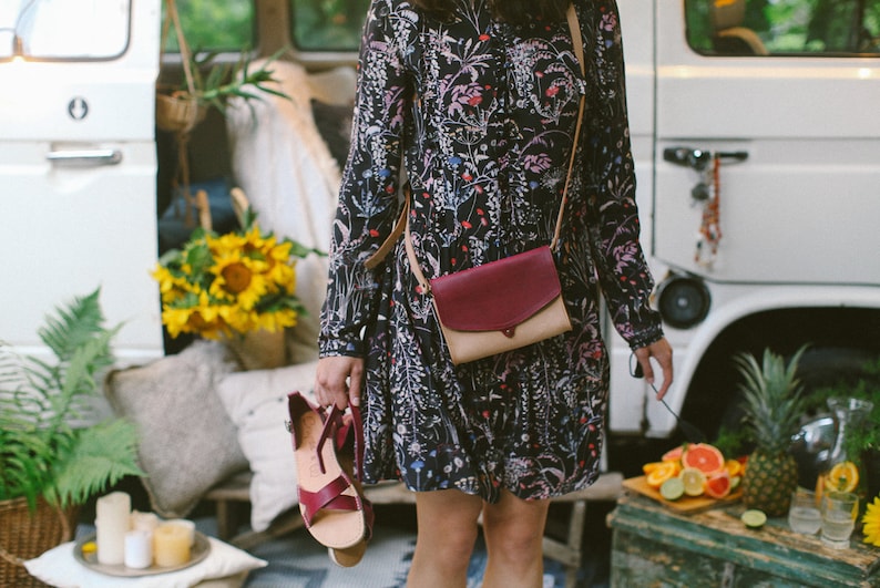 May include: A woman wearing a floral print dress and a burgundy and beige crossbody bag. The bag has a flap closure and a strap that goes over the shoulder. The woman is standing in front of a white van.