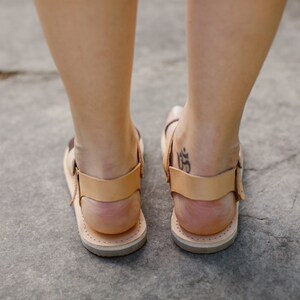 May include: A pair of brown leather sandals with a tan strap and buckle. The sandals are worn by a person with a tattoo on their ankle.