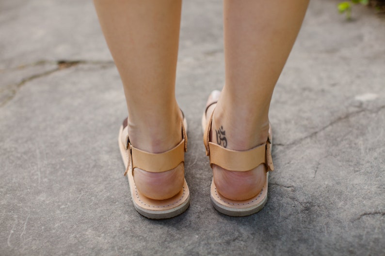 May include: A pair of brown leather sandals with a tan strap and buckle. The sandals are worn by a person with a tattoo on their ankle.