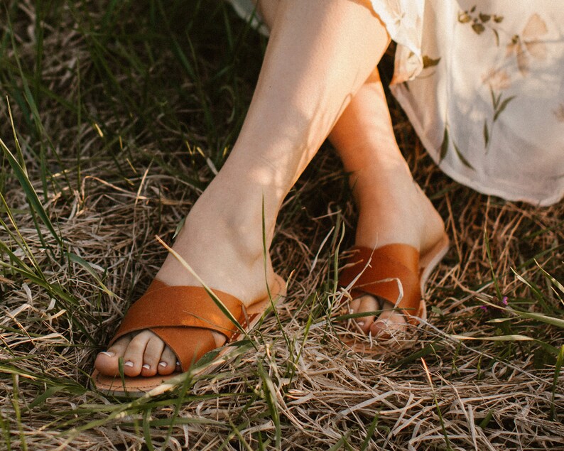 May include: A pair of brown leather sandals with a criss-cross strap design. The sandals are worn by a person sitting in a field of dry grass.