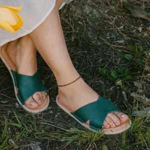 May include: A pair of dark green leather sandals with a criss-cross strap design. The sandals are worn by a person sitting on the ground with green grass and brown foliage.