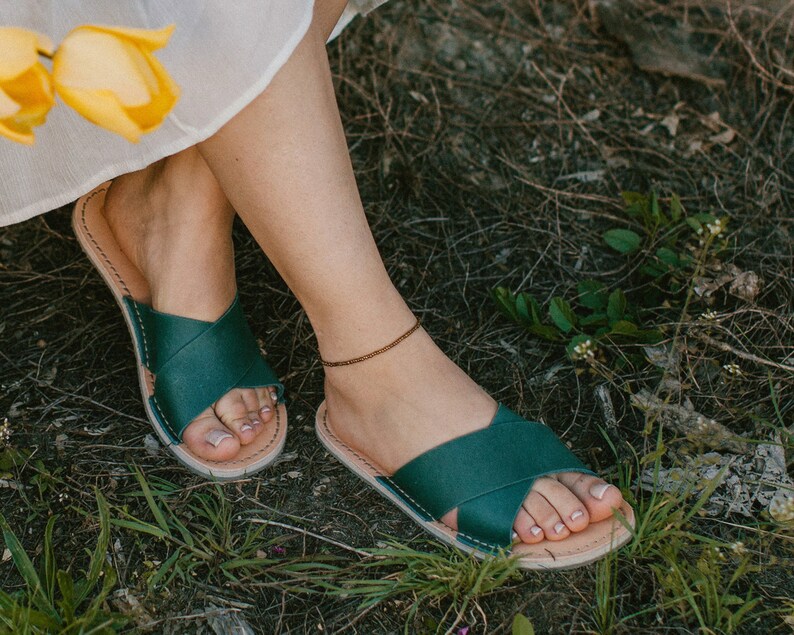 May include: A pair of dark green leather sandals with a criss-cross strap design. The sandals are worn by a person sitting on the ground with green grass and brown foliage.