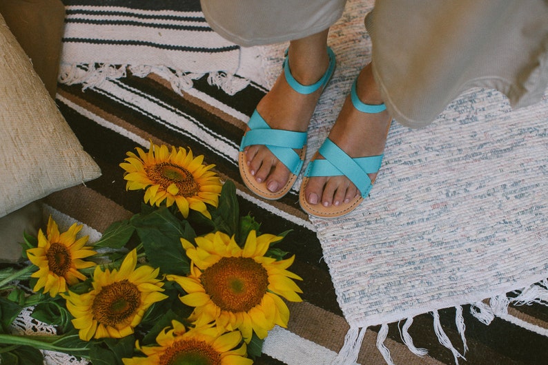 May include: A pair of light blue sandals with criss-cross straps. The sandals are on a white and brown striped rug with sunflowers in the foreground.