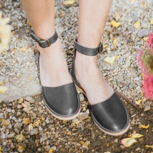 May include: A pair of gray leather ankle strap sandals with a closed toe. The sandals are on a gravel surface.
