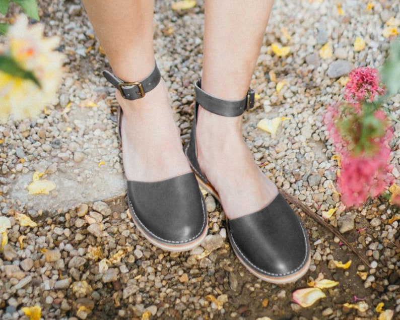 May include: A pair of gray leather ankle strap sandals with a closed toe. The sandals are on a gravel surface.