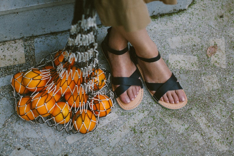 K&ouml;nnte beinhalten: Eine Netztasche gef&uuml;llt mit Orangen, mit einem Paar schwarzer Ledersandalen auf einem grauen Fliesenboden.