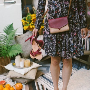 May include: A woman wearing a floral dress and sandals holds a red and beige leather crossbody bag. The bag has a flap closure and a strap that goes over the shoulder. The woman is standing on a striped rug.