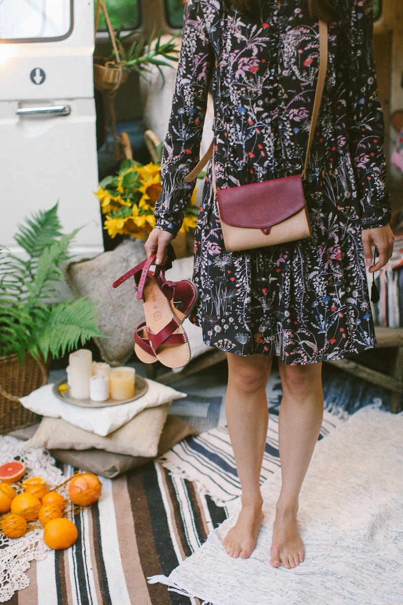 May include: A woman wearing a floral dress and sandals holds a red and beige leather crossbody bag. The bag has a flap closure and a strap that goes over the shoulder. The woman is standing on a striped rug.
