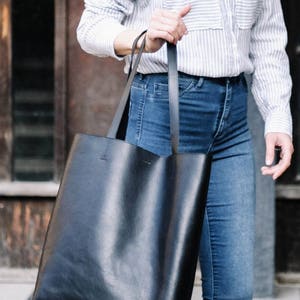 May include: A black leather tote bag with long straps. The bag is being held by a person wearing a striped blue and white shirt and blue jeans.