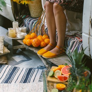 May include: A woman wearing a floral dress and yellow sandals sits on a bench with a bag of oranges. The bench is covered in a colorful striped blanket. There is a table with a pineapple, limes, and a glass pitcher in the foreground.