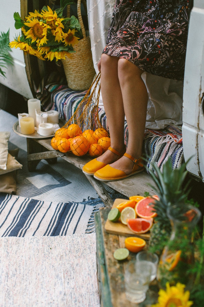 May include: A woman wearing a floral dress and yellow sandals sits on a bench with a bag of oranges. The bench is covered in a colorful striped blanket. There is a table with a pineapple, limes, and a glass pitcher in the foreground.