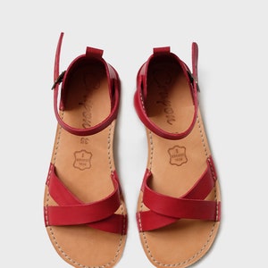 May include: A pair of red leather sandals with criss-cross straps and a buckle closure. The sandals have a brown leather insole and a brown leather sole. The sandals are made of real leather.