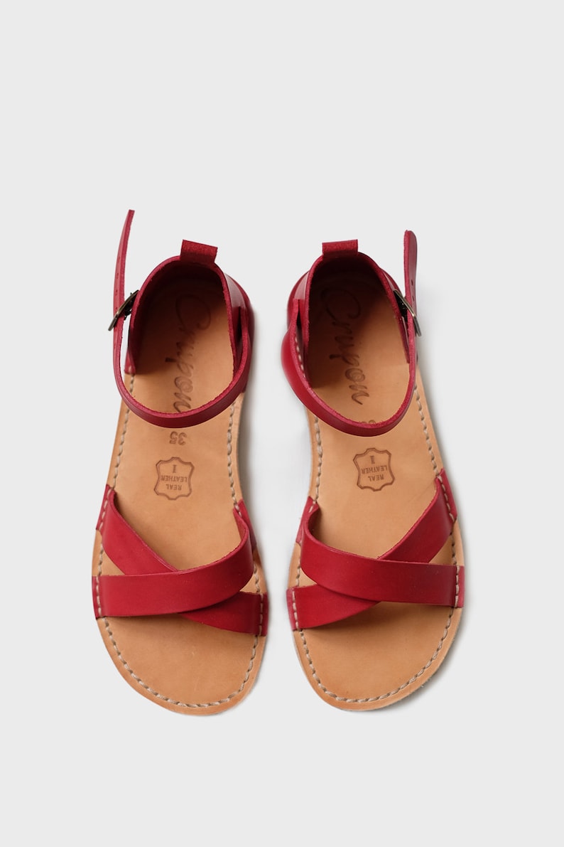 May include: A pair of red leather sandals with criss-cross straps and a buckle closure. The sandals have a brown leather insole and a brown leather sole. The sandals are made of real leather.