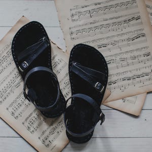 Peut inclure: Paire de sandales en cuir noir avec boucles, pos&eacute;es sur des partitions anciennes. Les sandales ont un design simple avec des lani&egrave;res et sont pr&eacute;sent&eacute;es sur un fond neutre.