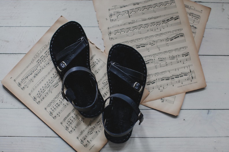 Peut inclure: Paire de sandales en cuir noir avec boucles, pos&eacute;es sur des partitions anciennes. Les sandales ont un design simple avec des lani&egrave;res et sont pr&eacute;sent&eacute;es sur un fond neutre.