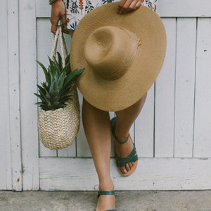 May include: A woman wearing a floral dress and a straw hat holds a woven basket with a pineapple inside. She is wearing teal sandals.
