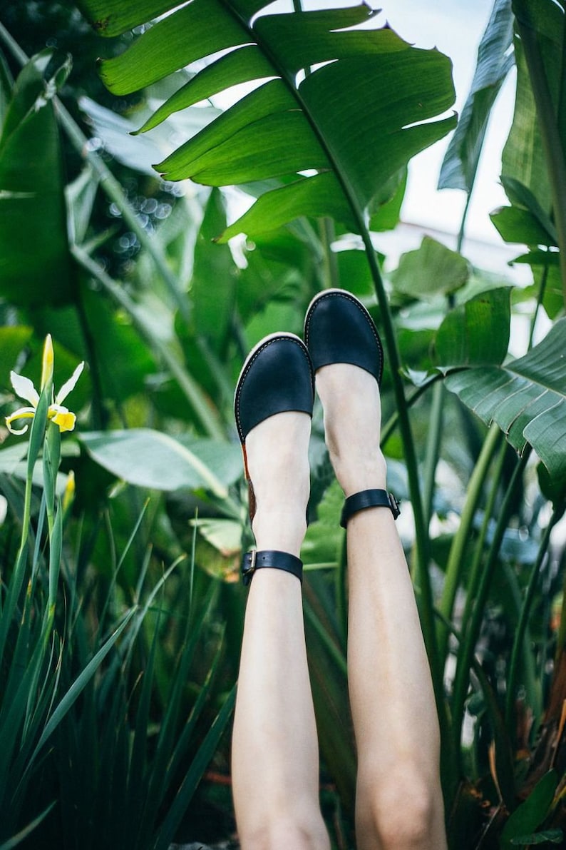 May include: A pair of black leather sandals with ankle straps, worn by a person with bare legs, against a backdrop of lush green foliage.