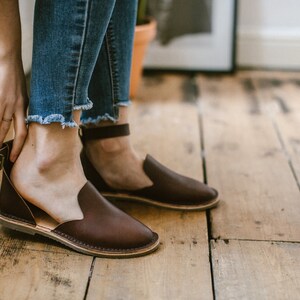 May include: Brown leather ankle strap sandals with a buckle closure. The sandals have a flat sole and are worn on a wooden floor.