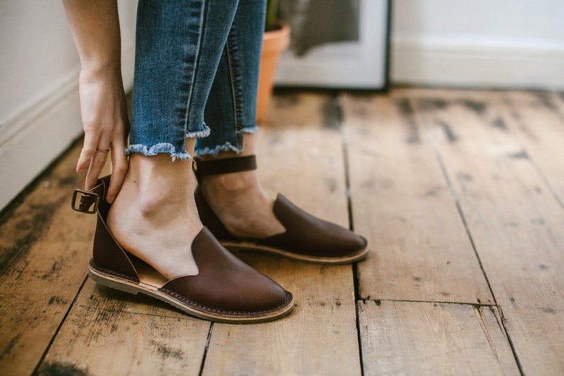 May include: Brown leather ankle strap sandals with a buckle closure. The sandals have a flat sole and are worn on a wooden floor.