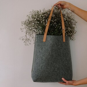 May include: A gray felt tote bag with brown leather straps. The bag is filled with white flowers.