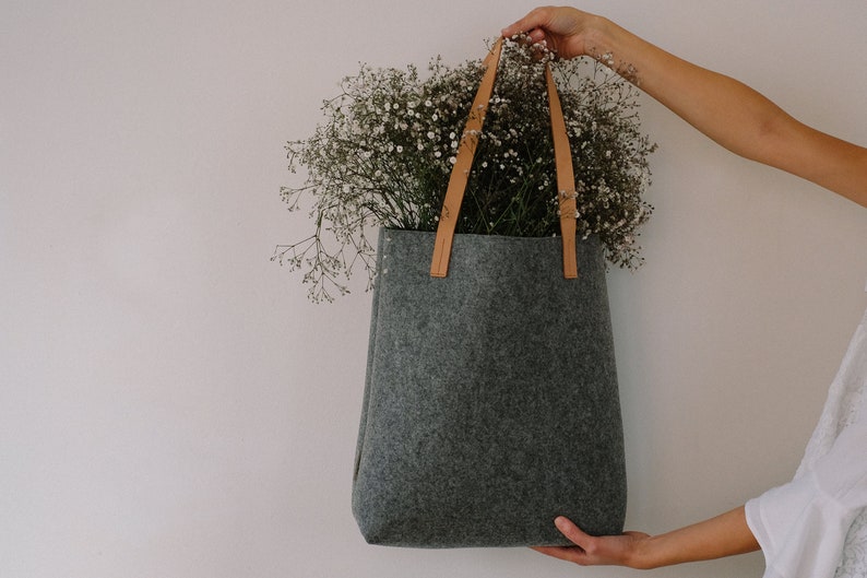 May include: A gray felt tote bag with brown leather straps. The bag is filled with white flowers.