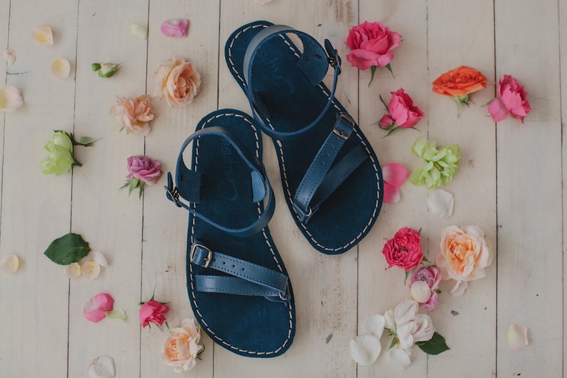 Peut inclure: Une paire de sandales en cuir bleu marine avec des coutures blanches et des fermetures &agrave; boucle. Les sandales sont entour&eacute;es de fleurs et de p&eacute;tales de roses color&eacute;es sur une surface en bois clair. Design minimaliste.