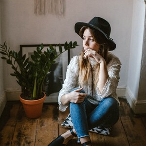 May include: A woman wearing a white sweater, blue jeans, and a black hat sits on a patterned cushion on a wooden floor. She is wearing black leather ankle strap shoes.