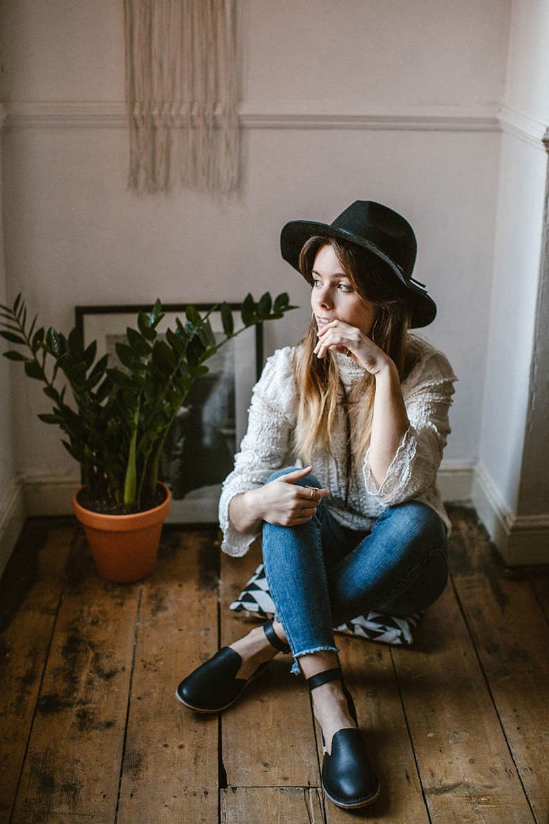 May include: A woman wearing a white sweater, blue jeans, and a black hat sits on a patterned cushion on a wooden floor. She is wearing black leather ankle strap shoes.