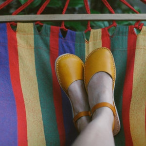 May include: A pair of yellow leather sandals with a strap across the foot, worn by a person relaxing in a colorful striped hammock.