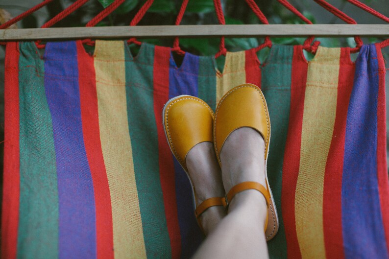 May include: A pair of yellow leather sandals with a strap across the foot, worn by a person relaxing in a colorful striped hammock.