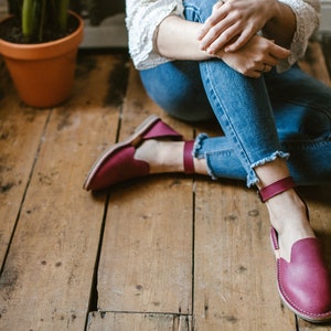 May include: A pair of burgundy leather ankle strap sandals with a closed toe and a flat sole. The sandals are worn by a person sitting on a wooden floor.
