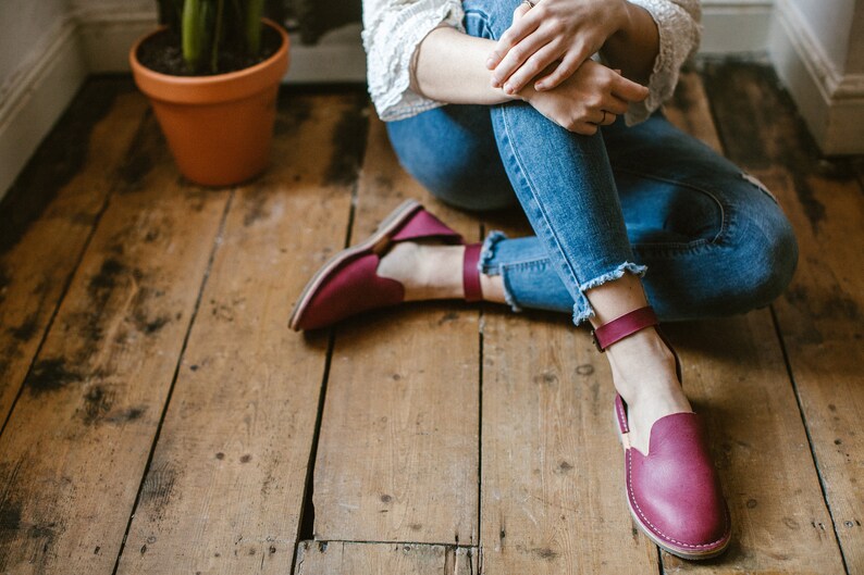 May include: A pair of burgundy leather ankle strap sandals with a closed toe and a flat sole. The sandals are worn by a person sitting on a wooden floor.