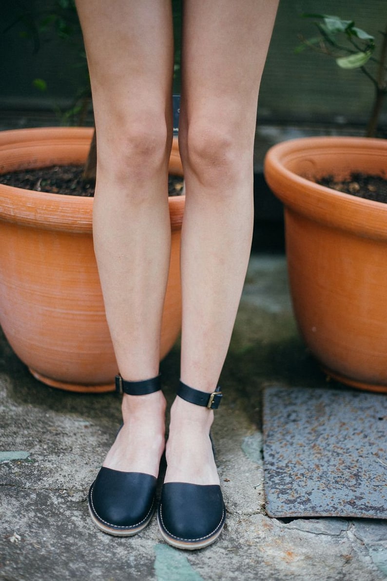 May include: A pair of black leather ankle strap sandals with gold buckles. The sandals are worn by a person standing on a concrete surface.