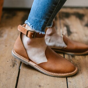 May include: Brown leather sandals with an ankle strap and buckle. The sandals have a cut-out design on the front and are worn on a wooden floor.