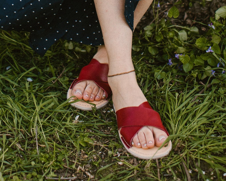 May include: Red leather sandals with a criss-cross strap design. The sandals are worn by a person sitting on grass.