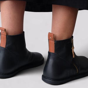 Pu&ograve; includere: Stivali da caviglia in pelle nera con un accento in pelle marrone sul retro e una cerniera laterale.